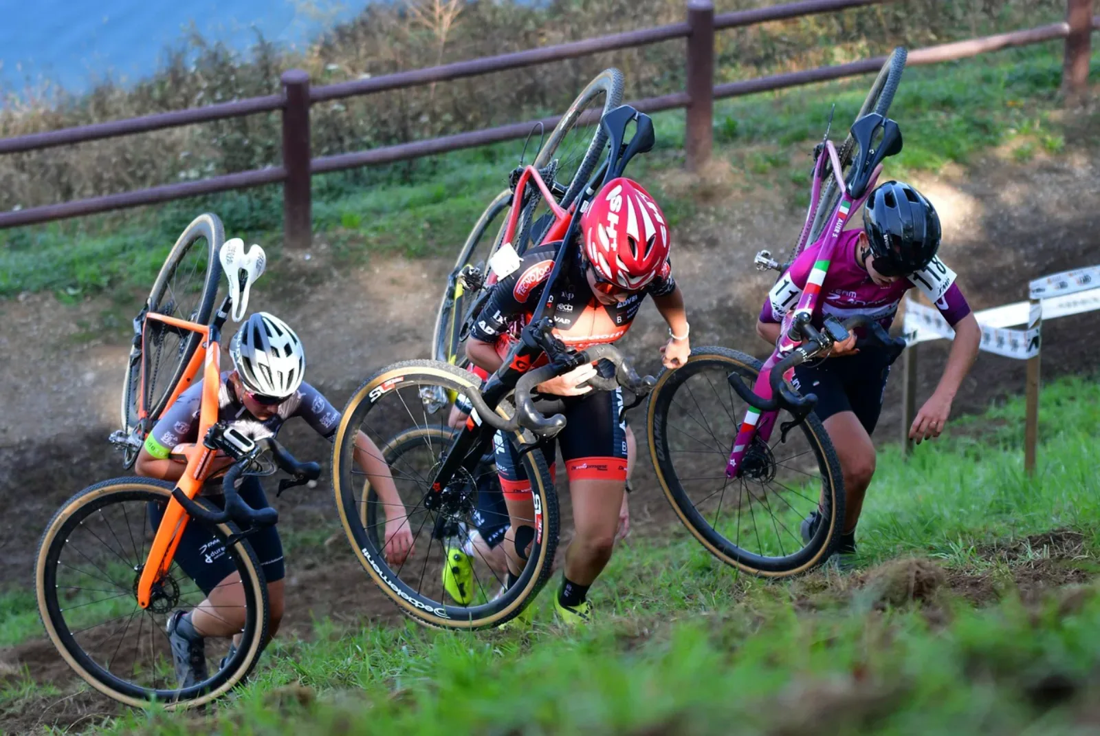 Trzy zawodniczki cyclocross niosą rowery na ramionach pod górę w trakcie wyścigu