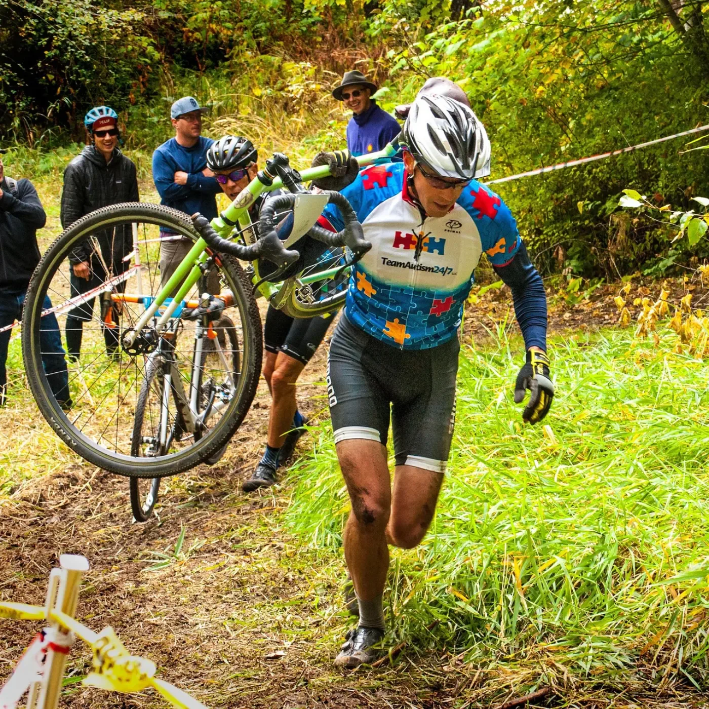 Mężczyzna w stroju kolarskim biegnie z rowerem cyclocross na ramieniu po leśnej trasie