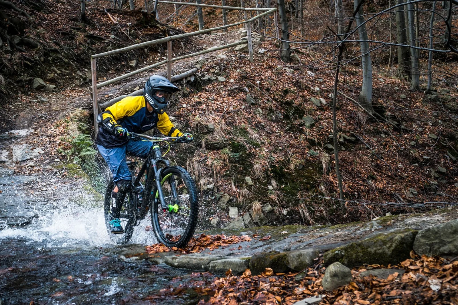 Rowerzysta enduro w żółtej kurtce i kasku pełnotwarzowym przejeżdża przez strumień — full suspension, jesienny las
