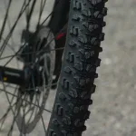 Zbliżenie na bieżnik opony MTB z agresywnymi kostkami i tarczę hamulcową w tle