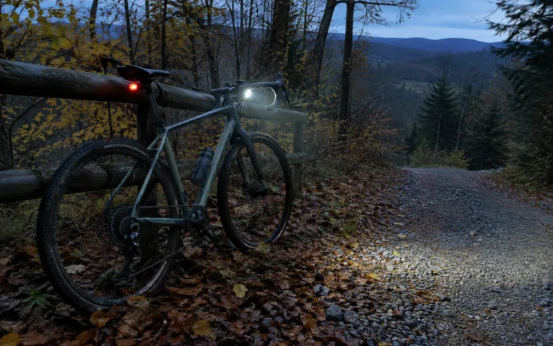 Rower gravel z włączoną lampką przednią i czerwoną lampką tylną na leśnej drodze o zmierzchu
